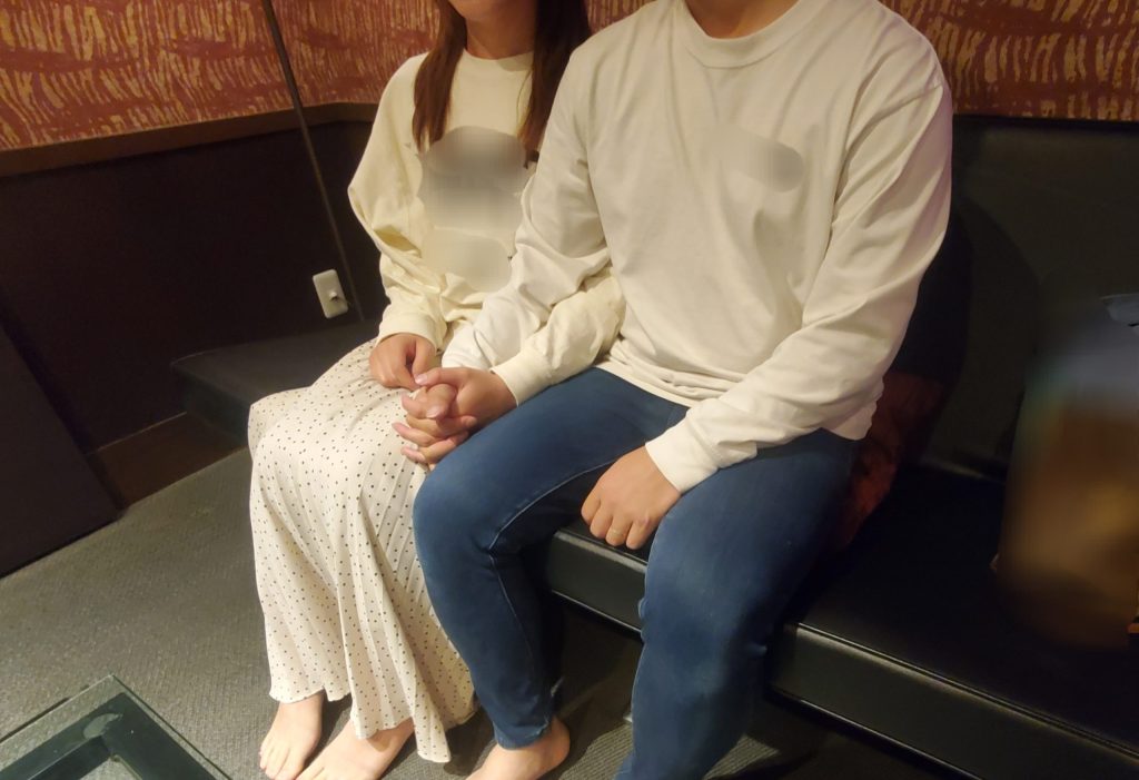 白い服を着た20代の若夫婦がソファに座り、手をつなぎながらNTR前の緊張感を漂わせている様子。寝取りの伝道師クロエによる寝取らせ企画。