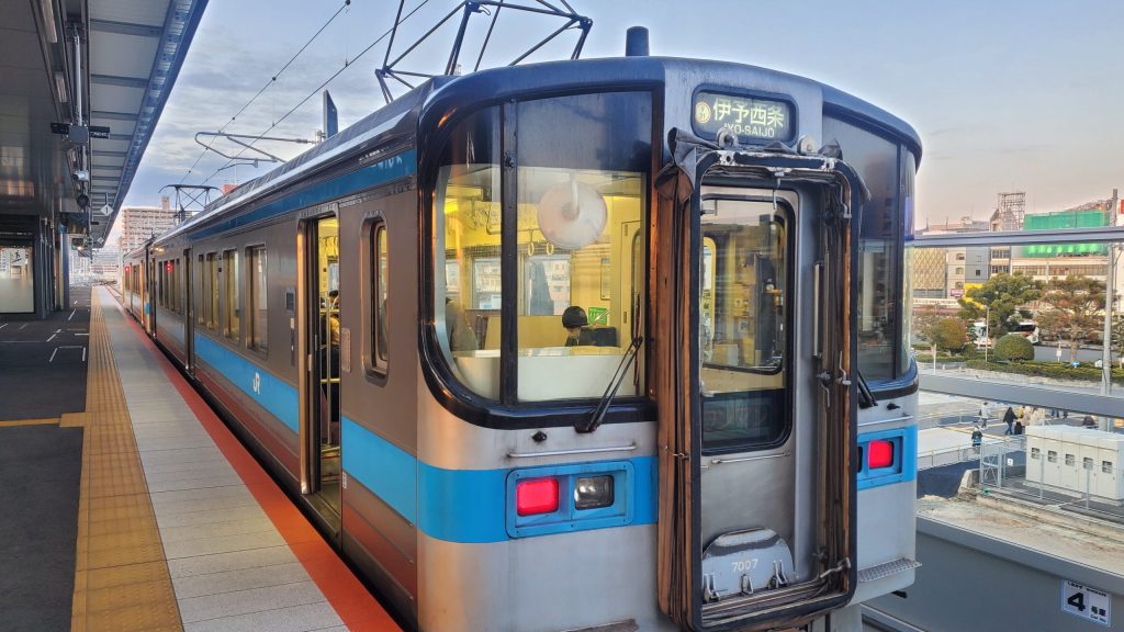 松山駅から今治へ向かうJR予讃線の列車画像。道後温泉観光を終え、愛媛県今治市のラブホテルで行われる寝取らせNTR体験へ移動する「ねとりっぷ愛媛編」の旅路を示す場面。
